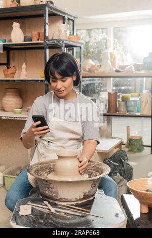 Smiling Brunette Craftswoman In Apron Making Clay Vase On Pottery Wheel