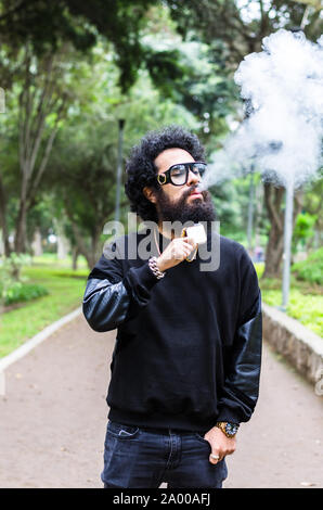 Vape. Young brutal man with large beard and fashionable haircut in sunglasses smoking an electronic cigarette in the city park. Steam cloud. Lifestyle. Stock Photo
