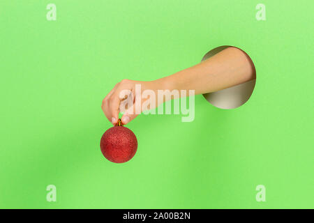 Kid holding red Christmas decoration Xmas tree bauble in hand through hole on light green background Stock Photo