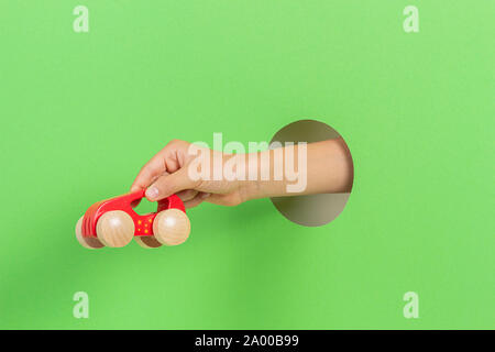 Kid holding red wooden toy car in hand through hole on light green background Stock Photo