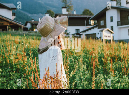 Beautiful romantic preteen girl in straw hat against the background of beautiful houses in the mountain, rural scene at sunset. View from behind, gold Stock Photo