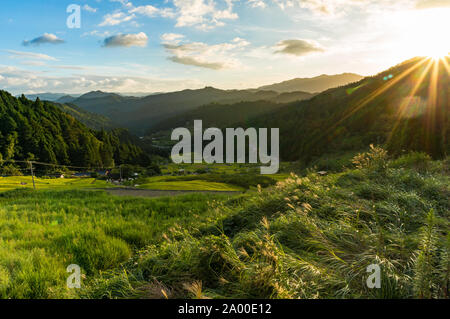 Sunset in the mountains with rice paddy fields and forest. Sun rays beaming over mountain peak with lens flare visible. Yotsuya, Aichi prefecture, Jap Stock Photo