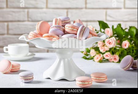 french pink and lavender macarons on a cake stand with beautiful bouquet of roses, cup of coffee and  brick wall at the background, close-up Stock Photo