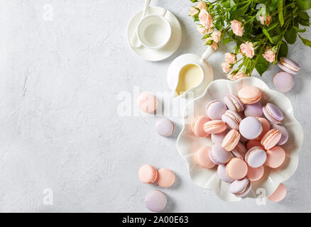 french macarons on a porcelain cake stand and some on a concrete table with bouquet of  fresh flowers and creamer, horizontal view, flat lay, empty sp Stock Photo