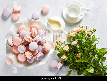 french macarons on a porcelain cake stand and some on a concrete table with bouquet of  fresh flowers and creamer, horizontal view, flat lay, close-up Stock Photo
