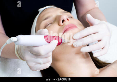 Cosmetologist,beautician in white gloves applying facial dermapen treatment on face of young girl customer in beauty salon. front side view Stock Photo