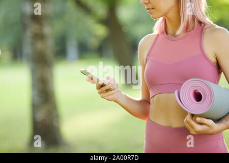 Close-up of young sporty woman in pink sports clothing holding exercise mat and looking at her mobile phone in her hand while standing outdoors Stock Photo