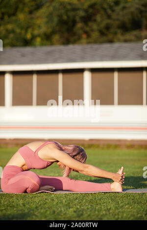 Young healthy woman in pink clothing sitting on exercise mat on green grass and stretching her body during training in the park Stock Photo
