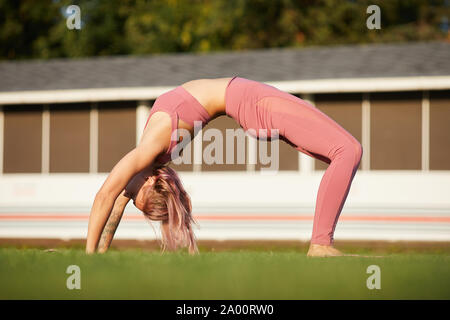 Young flexible woman in pink clothing doing stretching exercise on green grass in the park Stock Photo