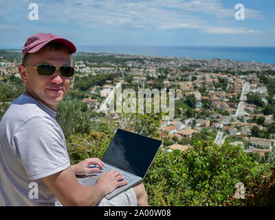A young male freelancer looks at the camera and sits on a mountain, with a beautiful landscape under it, a city by the sea, work in pleasure Stock Photo