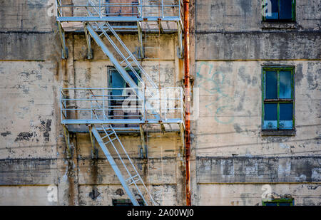 Plastered peeling wall of an old abandoned industrial multi-storey building with broken windows and a painted metal fire staircase and rusted pipes Stock Photo