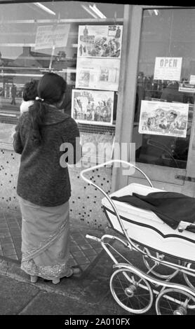 Southall, London 1972. A young mum looks at Indian movie posters in a shop window. After Idi Amin ordered 30,000 Asians out of Uganda with 90 days notice, those with British Passports headed to England. They were forced to leave their money, property and possessions behind. These images document life for some of the people as they settled in Southall, west of London. An insight into life 47 years ago for immigrants in the UK. Photo by Tony Henshaw Stock Photo
