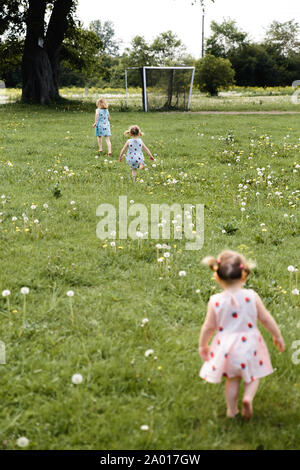 Mother having quality funny playing time with her baby girls at a park blowing dandelion - Young blonde hippie - Daughters wear similar dresses with Stock Photo