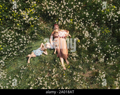 Aerial drone: Mother having quality funny playing time with her baby girls at a park blowing dandelion - Young blonde hippie - Daughters wear similar Stock Photo