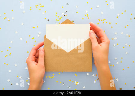 Female's hands holding kraft paper envelope letter with blank white card mockup over pastel blue background with golden confetti stars. Christmas, New Stock Photo