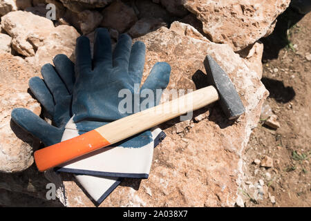 Close up of pair of protective gloves and masonry hammer on large stone boulder, with space for text on the right, on construction site. Salento, Pugl Stock Photo