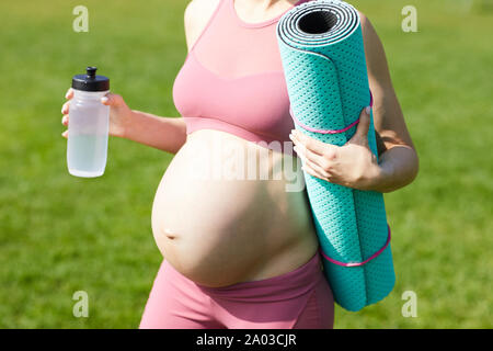 Close-up of pregnant woman in pink top holding exercise mat and bottle of water while standing outdoors after sports training Stock Photo