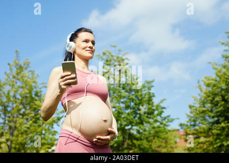 Young woman in pink sports clothing wearing headphones and using her mobile phone for listening to music while walking outdoors Stock Photo