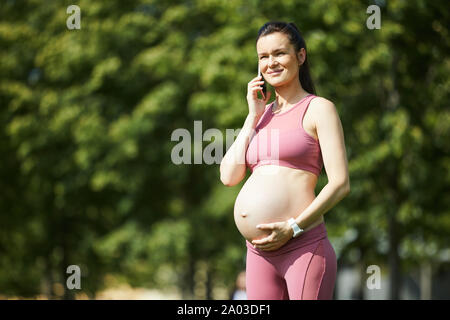 Young beautiful pregnant woman in pink clothing touching her belly and talking on mobile phone while standing in the park outdoors Stock Photo