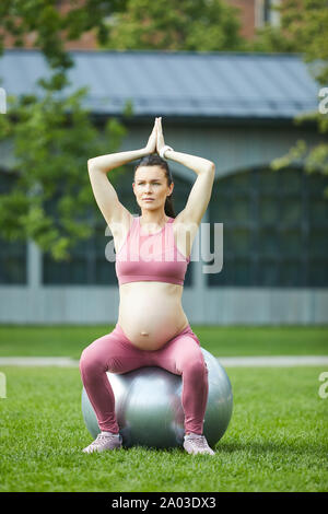 Young pregnant woman sitting on fitness ball raised her arms and doing exercise outdoors in the park Stock Photo