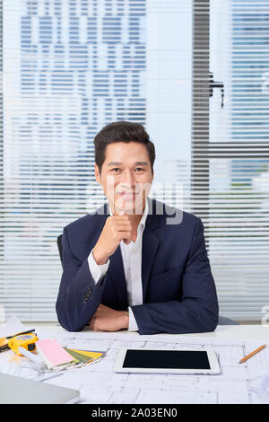 portrait of a nice smiling man, working at home on some project, he is sitting at a white table looking at camera, writing ideas with his laptop in fr Stock Photo