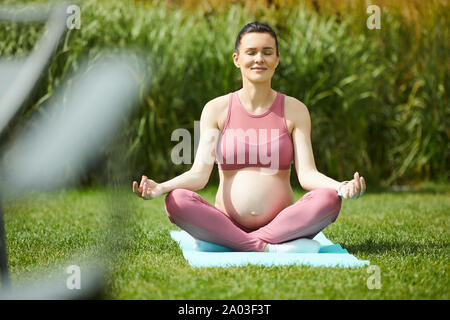 Portrait of healthy pregnant woman sitting on exercise mat in lotus position and doing yoga outdoors in the park Stock Photo
