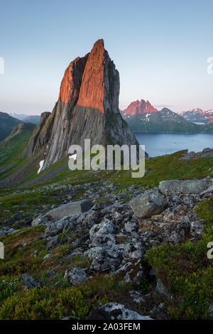 View from Mount Hesten on Iconic Mountain Segla in light of midnightsun in front of clear sky and mountain range in background, rocks and boulders in