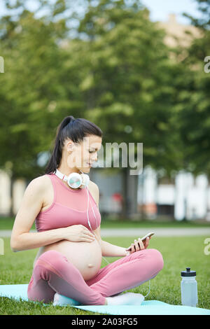 Young pregnant woman in pink clothing sitting on exercise mat and using mobile phone during sports training in the park Stock Photo