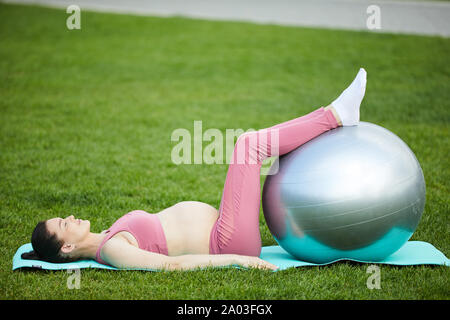 Young pregnant woman lying on exercise mat with her feet on fitness ball and exercising outdoors Stock Photo