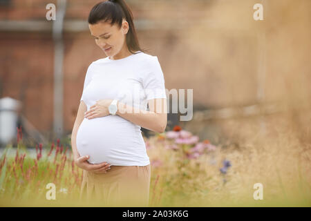 Young woman with dark hair embracing her pregnant belly and waiting for her baby while standing in the city Stock Photo