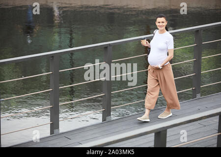 Portrait of young pregnant woman smiling at camera while drinking coffee and walking along the bridge outdoors Stock Photo