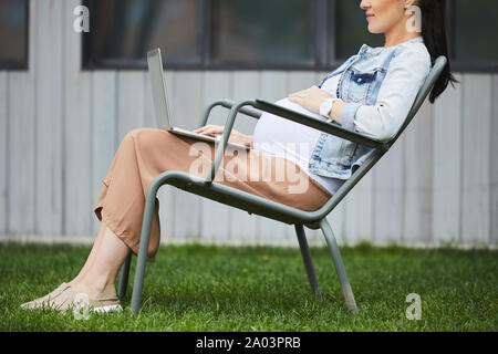 Close-up of young pregnant woman sitting on chair and communicating online using her laptop computer outdoors Stock Photo