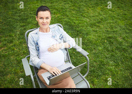 Portrait of beautiful pregnant woman sitting on chair with laptop computer on her knees and working online outdoors Stock Photo