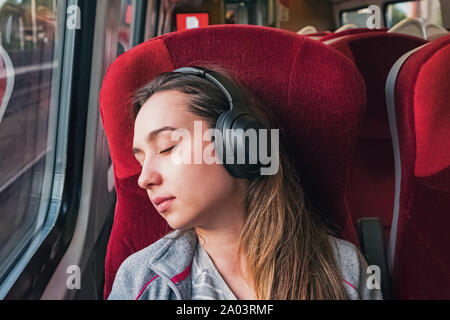 Young woman relaxing or sleeping while listening music in headphones sitting near the window in train Stock Photo