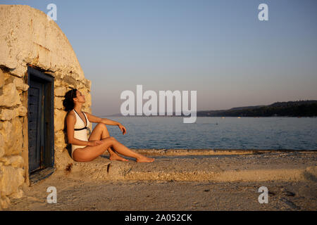 young beautiful woman in a swimsuit and sunglasses sits on a pier in Greece at sunset. girl enjoys the sun Stock Photo