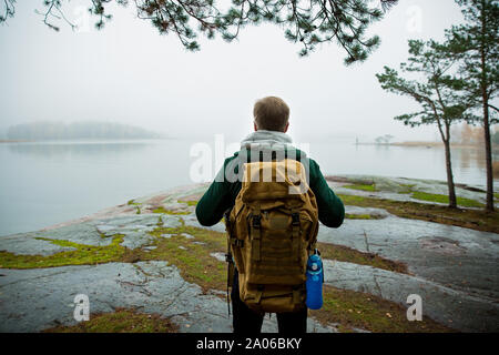 Mature man exploring Finland in the fall, looking into fog. Hiker with big backpack standing on mossy rock. Scandinavian landscape with misty sea and Stock Photo