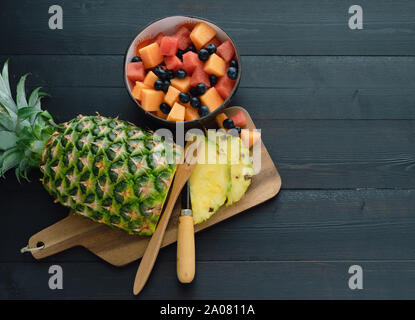 Sliced pineapple and bowl of fruits on black wooden background. Top view. Stock Photo