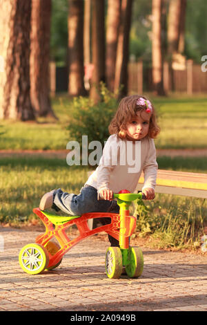Little toddler girl climbing on a bicycle in the sunlit summer park Stock Photo