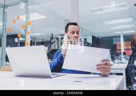 Male executive talking on mobile phone while using laptop at desk Stock Photo