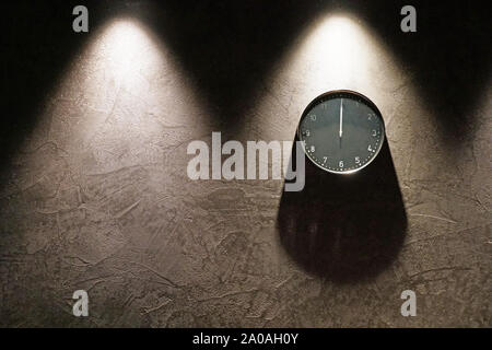 Black simple analog clock showing midnight hanging on the dark brown wall with copy space on the left. Three triangle shadows from above and one round Stock Photo