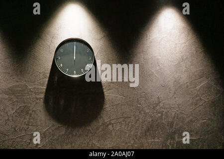 Black simple analog clock showing midnight hanging on the dark brown wall with copy space on the right. Three triangle shadows from above and one roun Stock Photo