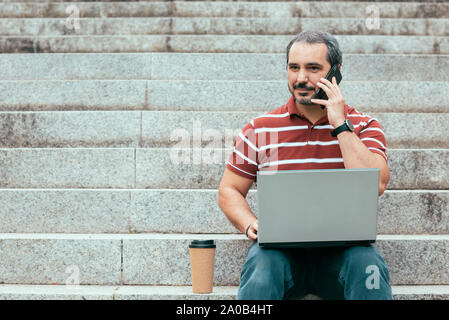 handsome middle-aged man holding laptop and talking on phone while sitting on stairs outside Stock Photo