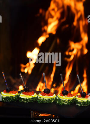 Cold appetizers, canape with red caviar, fire on background. Canape with olives, red caviar, lemon and dill. Restaurant dish concept. Delicious snacks Stock Photo