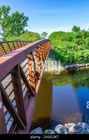 An arched footbridge with rust-colored metal railings crosses over a stream, connecting two sides of a pathway. Stock Photo