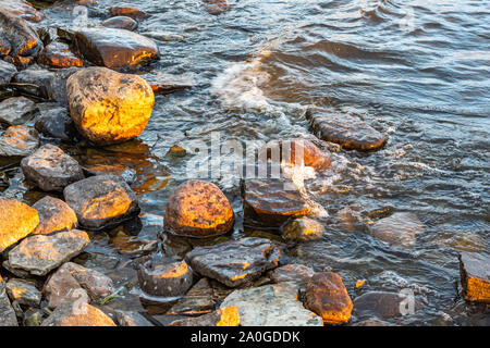 Small waves in a river roll into a rocky riverbank in the early morning light. Stock Photo