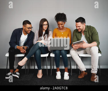Multiethnic friends sitting on chair obsessed with modern devices looking at laptop while waiting in queue Stock Photo