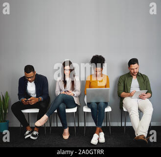 Multi-ethnic young people sitting in queue being addicted to social media using smartphones, digital tablet and laptop in office corridor Stock Photo