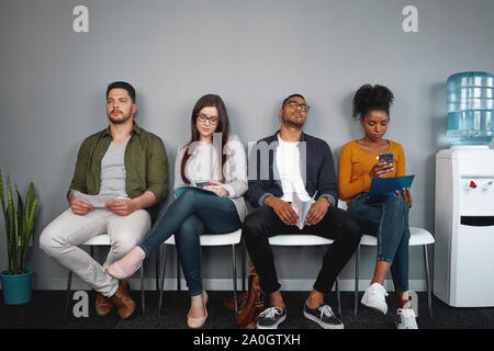 Diverse bored young applicant sitting on chair in row at waiting room for job interview in office - young group of real authentic people Stock Photo