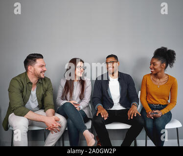 Multiracial happy young friends sitting relaxed and cheerful on chair in queue against gray wall - real authentic people being included and feeling a Stock Photo