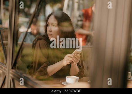 Charming brunette woman with long curly hair sitting at the window in cafe with mobile phone in hands Stock Photo